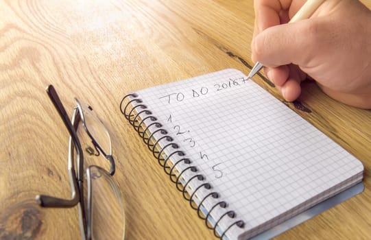 Man's hand writing the to do list for 2017, on a graph spiral notebook, placed on a wooden table with eyeglasses besides.