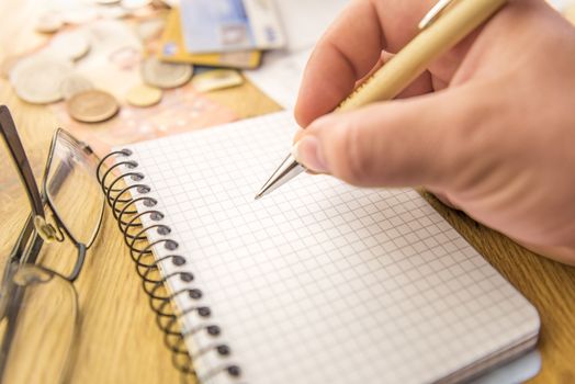 Close-up image with a blank math notebook and a man's hand holding a pen. In the background there are money, cards, bills and a pair of eyeglasses.