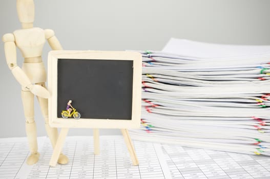 Wooden dummy holding empty blackboard with woman is cycling on finance account have blur pile overload document of report and receipt with colorful paperclip as background.