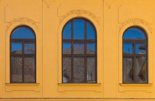 Ornate yellow facade of an old building with three windows with archs, reflecting a blue sky and a roof a the oposite building.