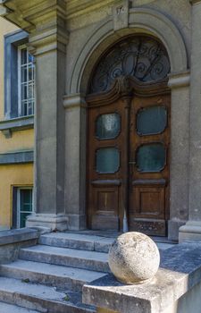 Side view on the entrance to an old building. Heavy wooden door with an arch. Stairs in a shadow and a stone ball enlightened by the sunlight.