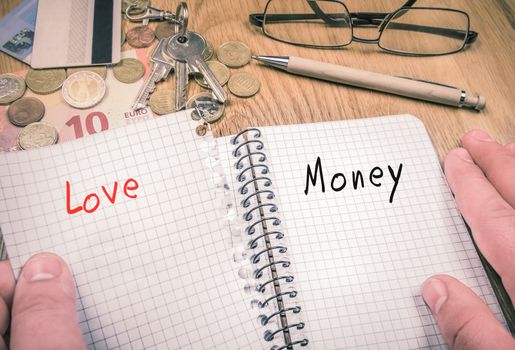 Man's hands ripping a sheet  with the word love, remaining the page with the word money in the notebook. There are financial elements and pen in the background.