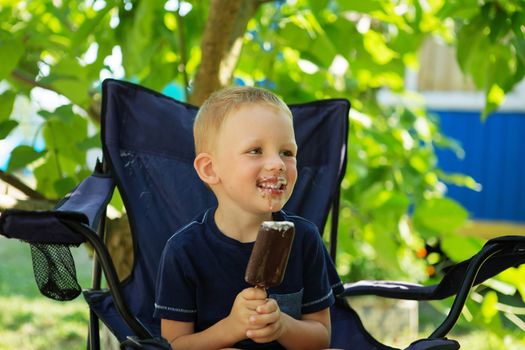 Adorable little boy eating ice cream pops in home's garden, outdoors. Happy blond kid eating chocolate ice cream bar