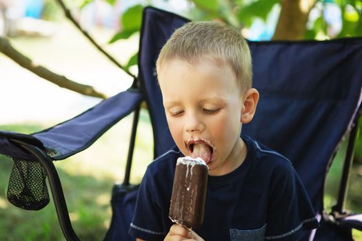 Adorable little boy eating ice cream pops in home's garden, outdoors. Happy blond kid eating chocolate ice cream bar