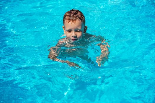 Happy kid playing in blue water of swimming pool on a tropical resort at the sea. Summer vacations concept. Cute boy swimming in pool water. Child splashing and having fun in swimming pool