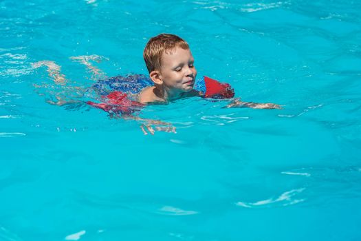 Happy kid playing in blue water of swimming pool. Little boy learning to swim. Summer vacations concept. Cute boy swimming in pool water. Child splashing and having fun in swimming pool