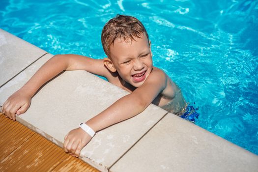 Happy kid playing in blue water of swimming pool. Little boy learning to swim. Summer vacations concept. Cute boy swimming in pool water. Child splashing and having fun in swimming pool