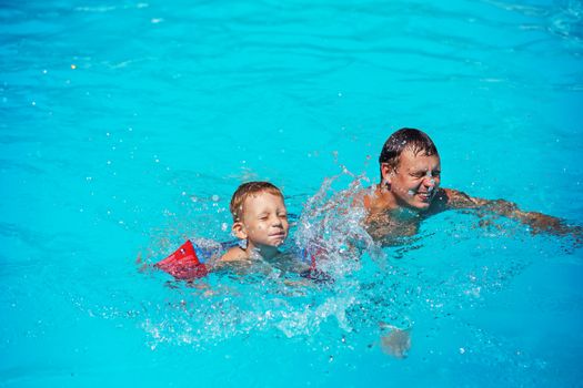 Happy boy enjoying with swimming and splashing with his father. Happy family playing in blue water of swimming pool. Kid learning to swim. Summer vacations concept. Dad and son having fun in swim pool