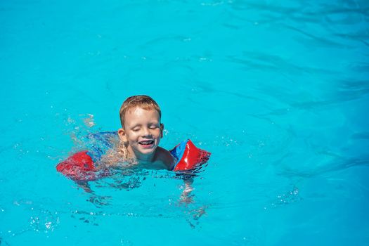 Happy kid playing in blue water of swimming pool. Little boy learning to swim. Summer vacations concept. Cute boy swimming in pool water. Child splashing and having fun in swimming pool