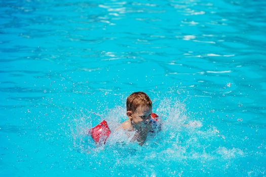 Happy kid playing in blue water of swimming pool. Little boy learning to swim. Summer vacations concept. Cute boy swimming in pool water. Child splashing and having fun in swimming pool