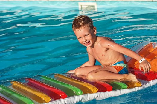 Little child on inflatable mattress in swim pool. Smiling boy playing and having fun in swimming pool with air mattress. Kid playing in water. Summer vacations concept. Boy swimming in pool water.