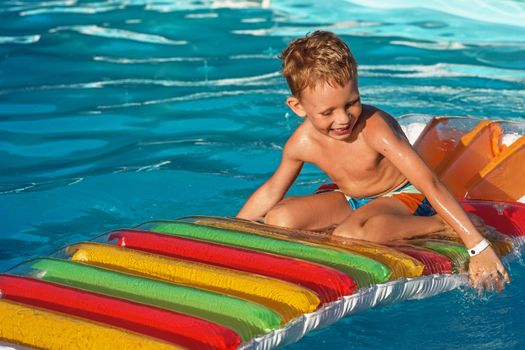 Little child on inflatable mattress in swim pool. Smiling boy playing and having fun in swimming pool with air mattress. Kid playing in water. Summer vacations concept. Boy swimming in pool water.