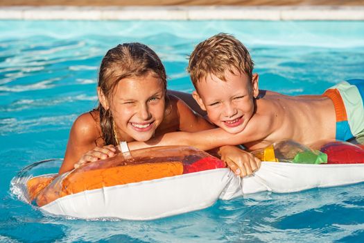 Little children on inflatable mattress in swim pool. Smiling kids playing and having fun in swimming pool with air mattress. Boy and girl playing in water. Summer vacations concept.