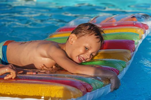 Little child on inflatable mattress in swim pool. Smiling boy playing and having fun in swimming pool with air mattress. Kid playing in water. Summer vacations concept. Boy swimming in pool water.