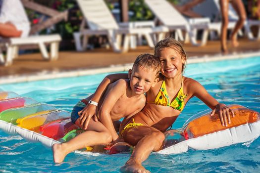 Little children on inflatable mattress in swimming pool. Smiling kids playing and having fun in swimming pool with air mattress. Boy and girl playing in water. Summer vacations concept.