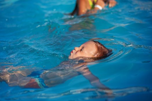 Happy kid playing in blue water of swimming pool. Little boy learning to swim. Summer vacations concept. Cute boy swimming in pool water. Child splashing and having fun in swimming pool