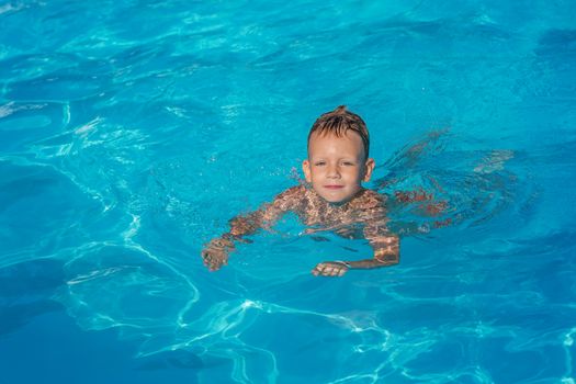 Happy kid playing in blue water of swimming pool. Little boy learning to swim. Summer vacations concept. Cute boy swimming in pool water. Child splashing and having fun in swimming pool