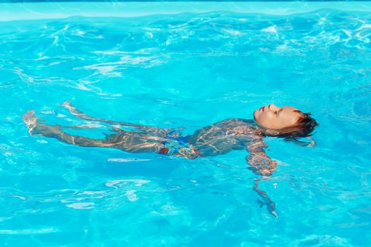 Happy kid playing in blue water of swimming pool. Little boy learning to swim. Summer vacations concept. Cute boy swimming in pool water. Child splashing and having fun in swimming pool