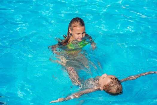 Happy kids playing in blue water of swimming pool. Children learn to swim on their backs. Summer vacations concept. Kids swimming in pool water. Boy and girl splashing and having fun in swimming pool