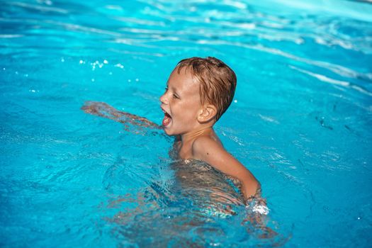 Happy kid playing in blue water of swimming pool. Little boy learning to swim. Summer vacations concept. Cute boy swimming in pool water. Child splashing and having fun in swimming pool