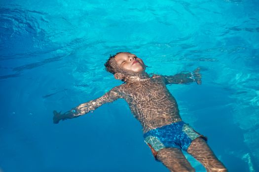 Happy kid playing in blue water of swimming pool. Little boy learning to swim. Summer vacations concept. Cute boy swimming in pool water. Child splashing and having fun in swimming pool