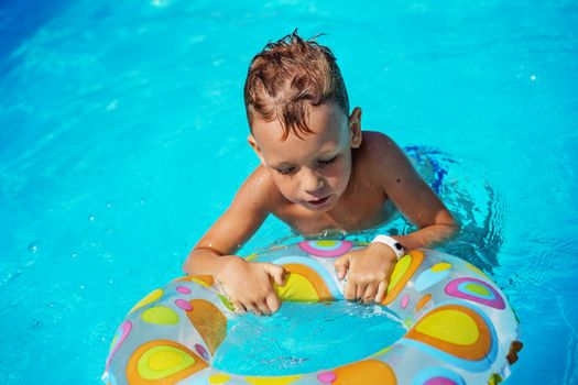 Happy kid playing in blue water of swimming pool. Little boy learning to swim with swim ring. Summer vacations concept. Cute boy swimming in pool water. Child splashing and having fun in swimming pool