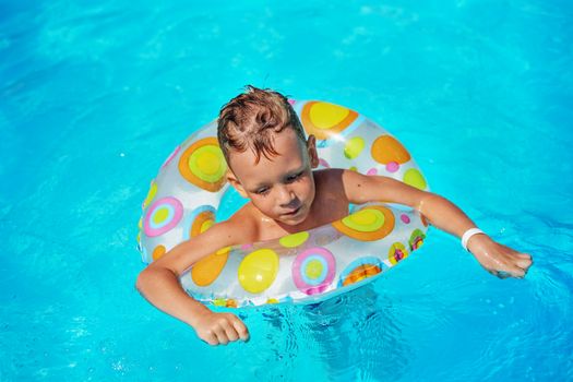 Happy kid playing in blue water of swimming pool. Little boy learning to swim. Summer vacations concept. Cute boy swimming in pool water with swim ring. Child splashing and having fun in swimming pool