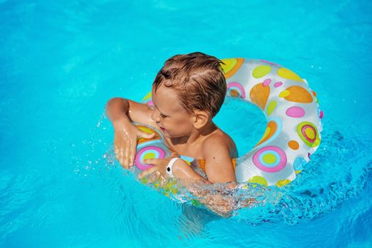 Happy kid playing in blue water of swimming pool. Little boy learning to swim with swim ring. Summer vacations concept. Cute boy swimming in pool water. Child splashing and having fun in swimming pool