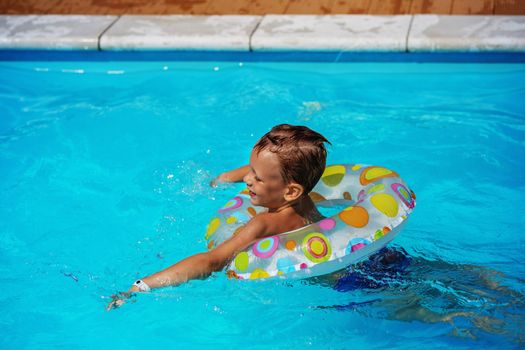 Happy kid playing in blue water of swimming pool with swim ring. Little boy learning to swim. Summer vacations concept. Cute boy swimming in pool water. Child splashing and having fun in swimming pool