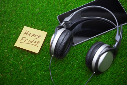 Happy Friday text on sticky note on a grass with headphones and digital tablet