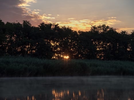 Sunrise in the river with reflex. Mirrored forest line on the river. Fantastic foggy river with fresh green grass in the sunny beams. Dramatic colorful scenery. 