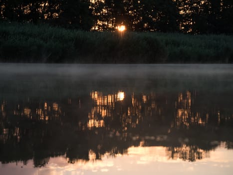 Sunrise in the river with reflex. Mirrored forest line on the river. Fantastic foggy river with fresh green grass in the sunny beams. Dramatic colorful scenery. 