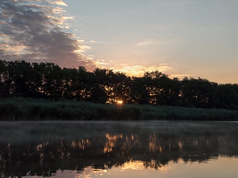 Sunrise in the river with reflex. Mirrored forest line on the river. Fantastic foggy river with fresh green grass in the sunny beams. Dramatic colorful scenery. 