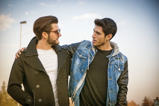 Two handsome casual trendy young men, 2 friends, in an urban park walking and chatting together