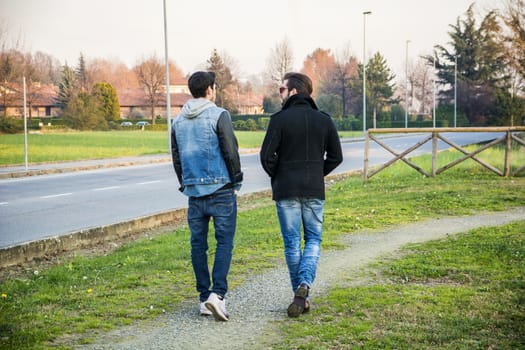 Two handsome casual trendy young men, 2 friends, in an urban park walking and chatting together