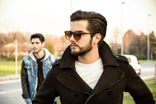 Two handsome casual trendy young men, 2 friends, in an urban park walking and chatting together