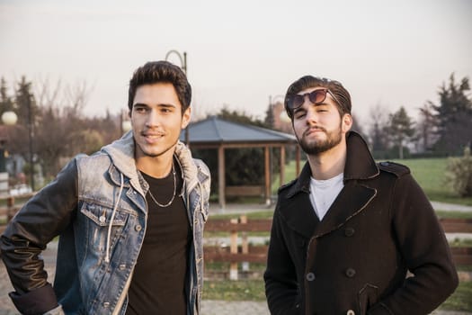 Two handsome casual trendy young men, 2 friends, in an urban park walking and chatting together