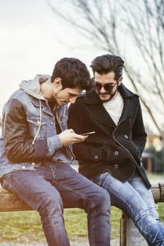 Two trendy casual young men sitting together on a bench outdoors reading an sms or text message, or looking at photos on a mobile phone