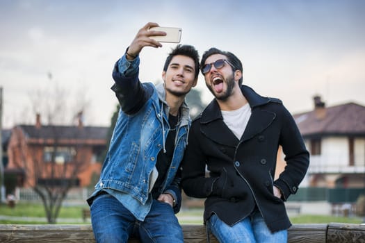 Two young men taking selfie while sitting on bench outdoors