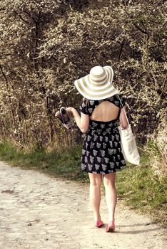Back view of cheerful fashionable woman in stylish hat and frock posing outdoor. Happy brunette girl with shoes and summer bag in hand