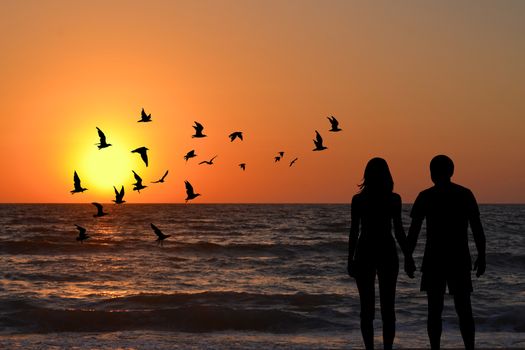 View of a full body of couple silhouettes holding hands looking at sunrise on the beach