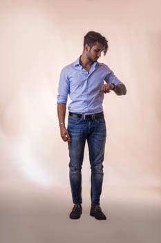 Portrait of brunette young man in light blue shirt and jeans, checking the time on wrist watch while standing in studio shot, isolated against white background. Full length photo