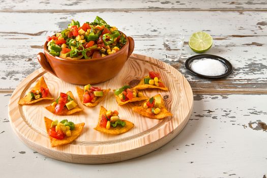 Nachos chips and vegetables in an earthenware bowl on a chopping board and tequila over a wooden table