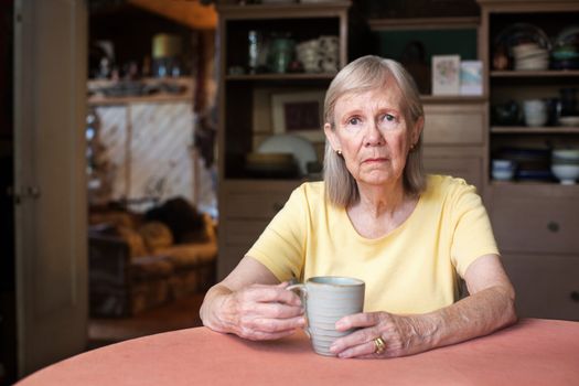 Senior woman in yellow shirt with depressed expression holding cup indoors