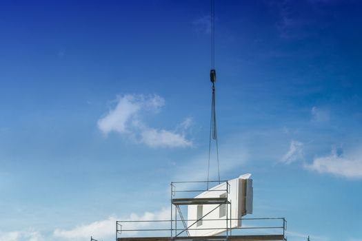 A wall part of a prefabricated house on a crane against blue sky.
