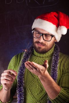 A young man with a beard in a green knitted sweater holding a Sparkler. The guy in glasses and a Santa hat welcomes the winter holidays. Old school