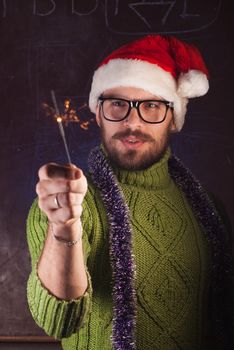 A young man with a beard in a green knitted sweater holding a Sparkler. The guy in glasses and a Santa hat welcomes the winter holidays. Old school