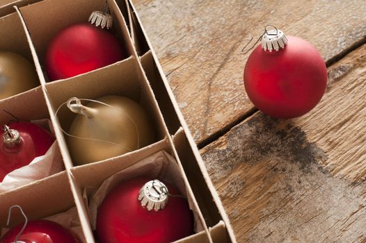 Red and gold Christmas baubles in a brown cardboard box with a single red ball outside on a rustic wooden table with copy space for a seasonal greeting