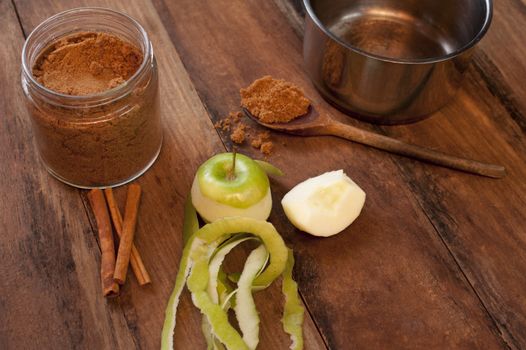 Fresh partly peeled apple, cinnamon sticks and powdered aromatic cinnamon spice in a spoon alongside a cup or small pot on a wooden kitchen counter