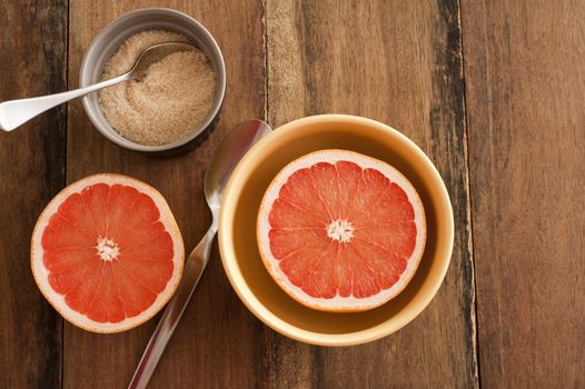 Fresh halved pink grapefruit served in a bowl for breakfast with caramelized sugar and spoons on a wooden table with copy space, overhead view